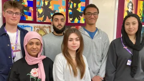 Alex Pope/BBC Six students, standing by a wall, with artwork on, looking at the camera. There are three girls and three men.
