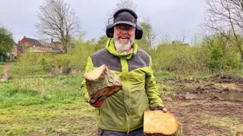 A man with a green and black jacket on. he has a black cap and ear defenders on. he is looking at the camera smiling with a chunk of would in each hand.