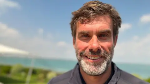 Iain Percy has medium length brown hair with a greying beard. He is wearing a zipped up navy jacket. The background is out of focus, but greenery and the sea can be made out in the distance.