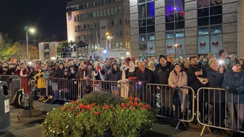 Shows a large group of people, some in fancy dress, looking up to the sky