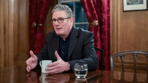 Keir Starmer gestures as he speaks while sitting in a room inside Downing Street wearing a dark shirt and suit with a mug and a glass of water on the table in front of him on Monday.