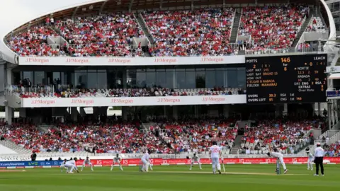 Getty Images A wide view of a cricket match in progress at Lord’s Cricket Ground, showing players on the pitch and a packed grandstand with spectators, sponsor signage, and a large electronic scoreboard in the background.