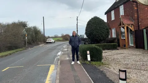 Nadira Tudor/BBC Terry Penton, wearing a navy bodywarmer, hoodie and trousers, stands with his hands in his pockets outside his brick home. On the left is a road with yellow bus stop markings painted on it. On the right is his house and a shingle drive.