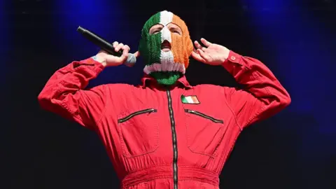 Getty Images A man wears a red boiler suit with a green white and orange flag on his left chest. He wears a balaclava that is the same colour as the flag. He puts both his hands up to his ears. His right hand holds a black microphone.
