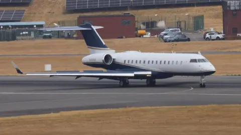 SWNS A private plane taxis along the tarmac at Birmingham Airport. Picture taken from a side angle, with the planes windows and one wing visible. It is a white aircraft with navy blue wings and tail