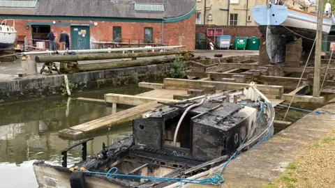 A burnt-out boat in front of a boat's body at the the end of a harbour. In the background you can see men stood near a red-brick building with green doors.