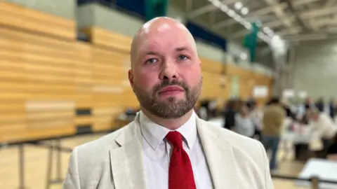 Close view of a man in a light-coloured suit and red tie standing in a large indoor hall during an election count, with tables, ballot boxes and people working in the background