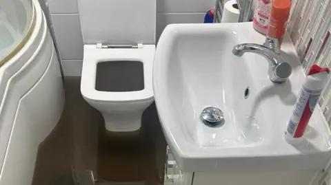 Barry Froggatt A flooded bathroom. We can see brown water on the floor and in the toilet seat. We can see a sink and the edge of a bath. 
