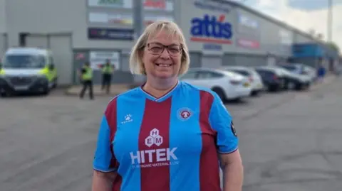 A woman with short blonde hair and glasses wearing a Scunthorpe United top and standing in front of Attis Arena smiling.