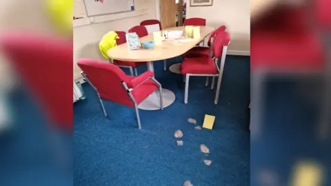 Wiltshire Police An office-type room with a set of table and chairs. On it, a lunch box and bowl has been place with folded yellow paper to look like crime scene labels. On the floor, muddy footprints have been purposely put in place.