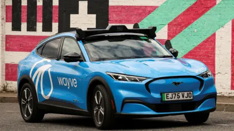 Reuters A blue car with Wayve branding and a roof‑mounted sensor unit, parked in front of a brightly painted mural wall