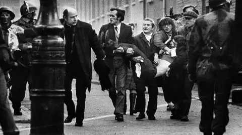 Pacemaker A picture taken on 30 January 1972 - Bloody Sunday. A group of men are carrying a lifeless body - they are accompanied by a priest waving a wife handkerchief. There are soldiers at the side of the picture and in the foreground.