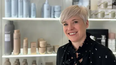 Kerry Larcher, a woman with short blonde hair wearing a black dress, in front of shelves containing hair products. She is smiling at the camera