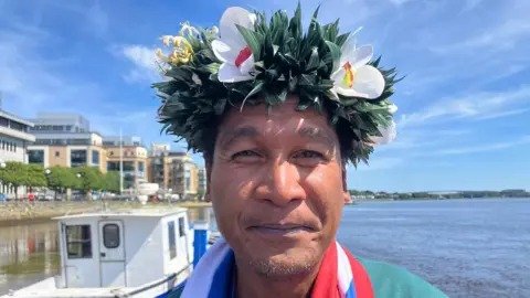 Willison Yarofaitir, wearing a traditional Micronesian head dress, stands on the water's edge. He is wearing a green t-shirt and is smiling, a boat can be seen over his right shoulder.