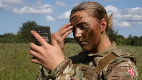 Reuters A soldier in the Danish Royal Life Guards applies camouflage paint during a training exercise in Hovelte, Denmark, on 11 June 2025.