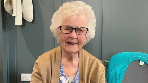 A photo showing the head and shoulders of an elderly white woman, She is wearing glasses and has white hair. She is sat down in front of a dark wall