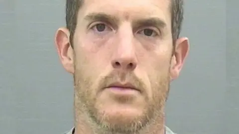 Timothy Brehmer, a man with stubble and short dark hair, looks into the camera for a police mugshot.