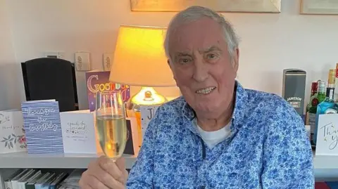 BBC DJ Johnnie Walker seen inside his home, against a backdrop of thank you and goodbye cards. He is wearing a blue floral shirt and smiling as he holds a glass of champagne in his right hand.