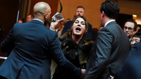 Lidia Thorpe, a dark haired woman wearing a possum fur cloak, is surrounded by men in suits as she shouts in the Canberra Great Hall