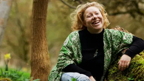 Fotografi av Josephine Slade Athena Aperta sitter på en tømmerstokk i skogen. Det er andre trær rundt den. Hun smiler og har på seg en grønn skjorte over en svart topp.