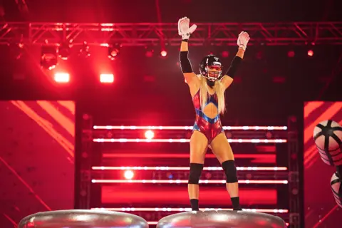 Credit BBC / James Stack / © Hungry Bear Media Ltd A woman with long blonde hair, wearing a black helmet and a red and blue Gladiators outfit, stands on a podium during the show with her arms in the air.