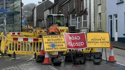 The picture shows a residential street that has been dug up for roadworks. Yellow safety barriers line the work area, and a construction worker in an orange high‑visibility jacket is standing beside a small digger parked in the middle of the road. Several signs are placed in front of the barriers. One warns that the road will be closed from early January to February 2026. Another sign reads “Road Closed”, and another directs drivers towards access for a medical centre.