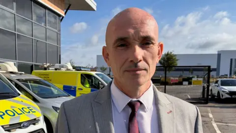 Linsey Smith/BBC Detective Superintendent Al Curtis is dressed in a grey suit with a burgundy tie. He is stood in front of three police vehicles in a car park. He is looking at the camera with a sombre expression.