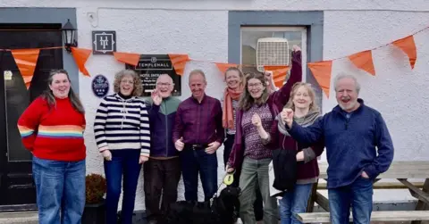 MCHP Eight people - five women and three men - lined up outside the pub with small orange triangular flags strewn above their heads, punch the with air with delight.