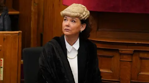 PA Media Dorothy Bain KC mengenakan wig putih dan jubah hitam di ruang sidang kayu. Dia memiliki rambut hitam dan mengenakan atasan putih.