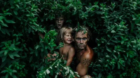 Maria Vatne Nik Payne, who is topless, peers from inside a leafy bush with his young sons on his knee