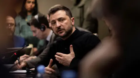 Ukrainian President Volodymyr Zelensky, wearing a black top, looks directly into the camera. Member of the press can be seen in the background, and there is a microphone on the table in front of him