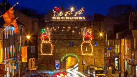 Alnwick Christmas Lights Committee Bondgate Tower in Alnwick, which is a medieval structure consisting of a large gatehouse with a road running through a narrow arch in the middle, is covered with Christmas lights with Santa at the very top.