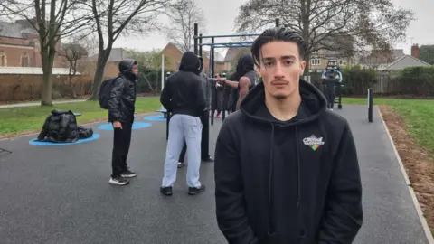 LDRS Theo Dopson wearing a dark hoodie stands in the foreground of an outdoor fitness area. Behind this person, several others are gathered around metal workout bars. The ground is covered with rubber safety surfacing, and leafless trees and nearby houses can be seen in the background on an overcast day.