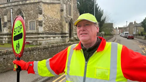 Matthew Crossman standing in a road near a church with his arms outstretched to stop traffic, holding his "stop" sign. He has a hi-vis yellow and orange jacket on over a dark green fleece top. A bit of dark hair is visible under his yellow cap.