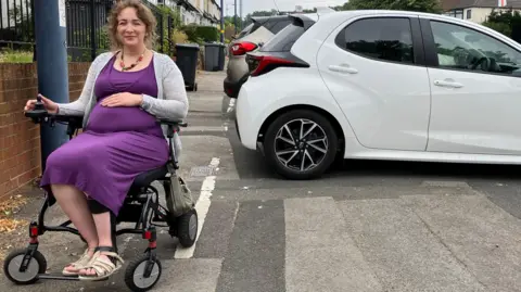 Hana, a woman with brown, shoulder-length hair, sits in a wheelchair, with one hand on her lap. She wears a purple dress, a grey cardigan and grey sandals. She is positioned just to the left of the white line on the street - behind her are wheelie bins, while cars occupy the pavement on the other side of the street.
