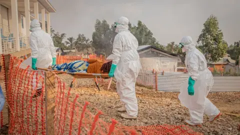 Getty Images A woman is carried on a stretcher by people wearing white hazmat suits through a dry field with sheds and fencing in the background 