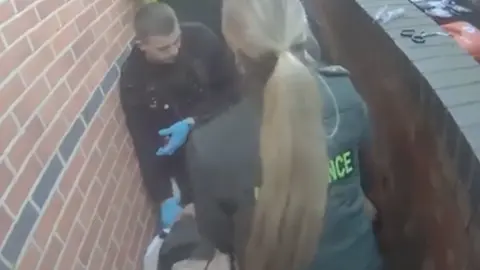 A grainy photo from a video showing a narrow alleyway with a police officer with cropped hair wearing blue plastic gloves leaning over someone. An ambulance worker with a long, fair ponytail is also standing over the man, with her back to the camera.