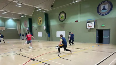 A player wearing a skull cap and a red Man Utd football top is about to shoot as the goalkeeper runs out to close him down. Players are doing a training drill around them. 