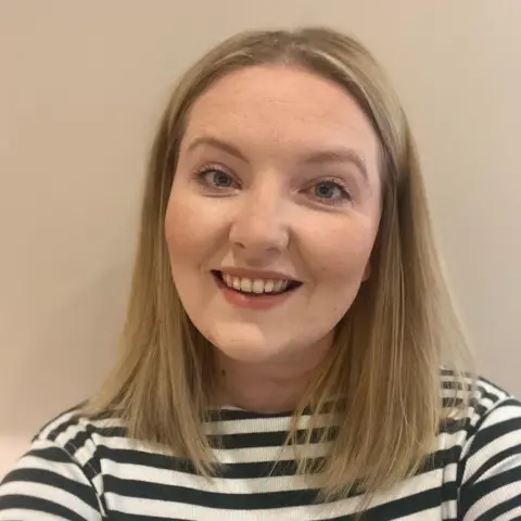 Charlotte Schofield A blonde woman looking down the barrel of the camera smiling. It is a head and shoulders shot and she wears a black and white striped top. 