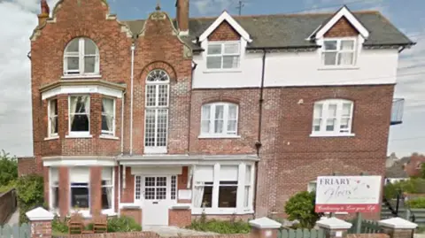 Friary House is a detached, three-storey, brick building behind a low wooden fence.