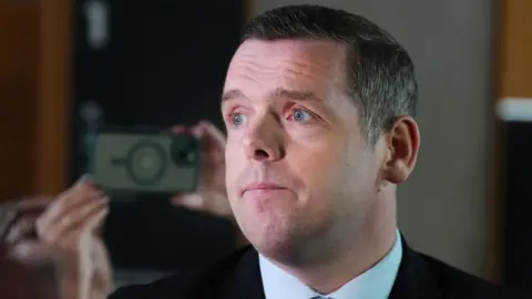 PA Douglas Ross, who has short dark hair, in a close-up shot with a flash illuminating his face. He is wearing a dark suit and white shirt, with a phone being held up behind him to take a photograph. 