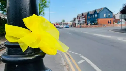 A close up of a yellow ribbon tied to a black lamppost with village buildings in the distance