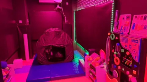 Hull City Council A room with reddish-coloured walls, a tent and puzzle games. There is a pink shelf unit and a blue mat on the floor.