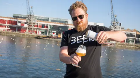 Bristol Craft Beer Festival A man with ginger hair and a beard, wearing a black T-shirt with white writing which says 'beer', is pouring a glass of beer from a can. He is standing in front of Bristol's harbour, with a museum and historic cranes in the background.