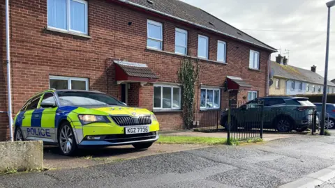 A police car is parked outside a house. The car is is blue and yellow and has the word police printed on the front and the side. 