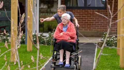 Healthcare Management Trust An elderly woman in a wheelchair being pushed by a younger woman