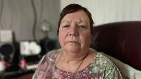 Jo Makel/BBC Sue, a 69-year-old woman with short, brown hair, and a fringe swept to one side, looks straight at the camera. She is sat on a sofa.  The photo is just of her head and shoulders. She is wearing a purple and green patterned top. Her glasses hang on a chain around her neck.