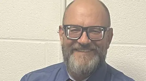Chris Parry Close up of Chris smiling whilst looking at the camera. He stands in front of a white bricked wall and wears a blue shirt and black glasses. He has short grey hair and a grey beard. 