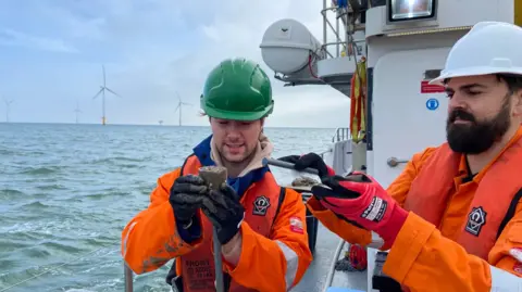 Two researchers dressed in orange waterproofs, the man on the left wearing a green hard had and the man on the right wearing a white hard hat, each lifting up and looking at seabed samples, on a white boat with the sea and windfarm turbines behind them 