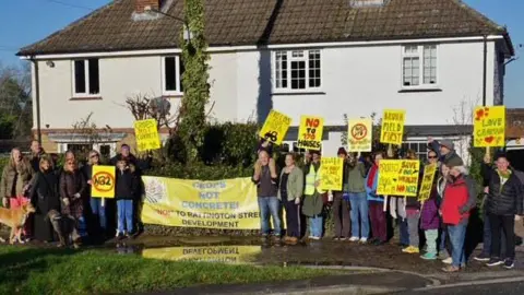 A group of people holding up placards and signs opposing a housing proposal on the edge of a village. 